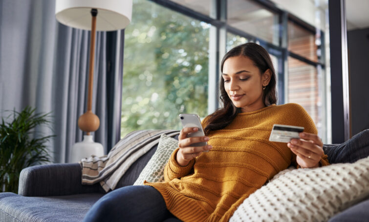 Shot of a beautiful young woman using her cellphone and credit card while relaxing on a couch at home