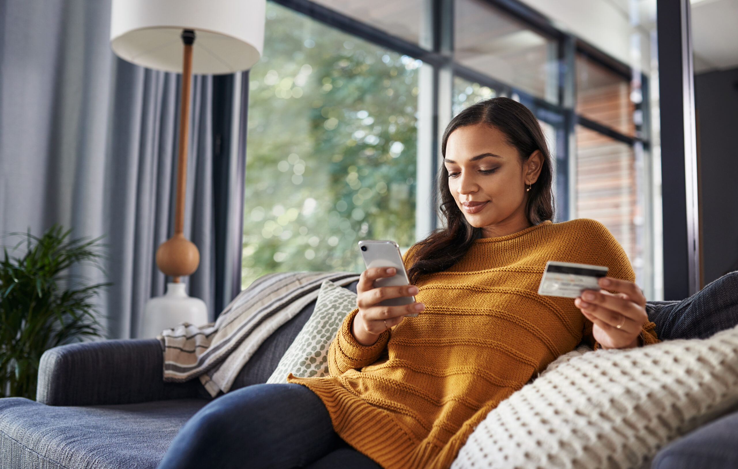 Shot of a beautiful young woman using her cellphone and credit card while relaxing on a couch at home
